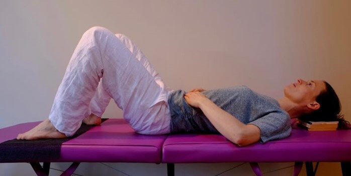  person laying on back, knees bent, hands on tummy, head resting on books on treatment table