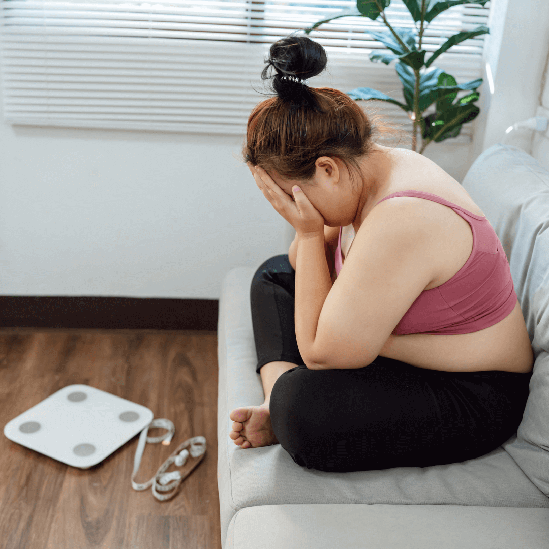 Woman sitting on couch with head in hands, frustrated with weight loss efforts, highlighting the benefits of hypnotherapy and virtual gastric band for effective weight management.