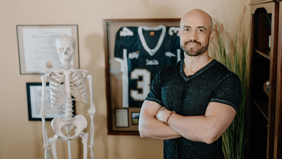 Dr. Ryan Johnson standing in his office with arms crossed, next to anatomical skeleton and framed football jersey.
